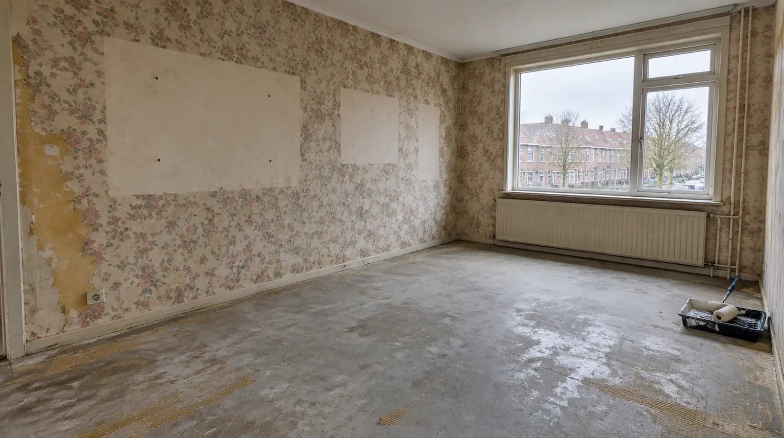 Lege kamer met half verwijderd bloemetjesbehang, lichte plekken waar schilderijen hingen en lijmresten op de betonvloer