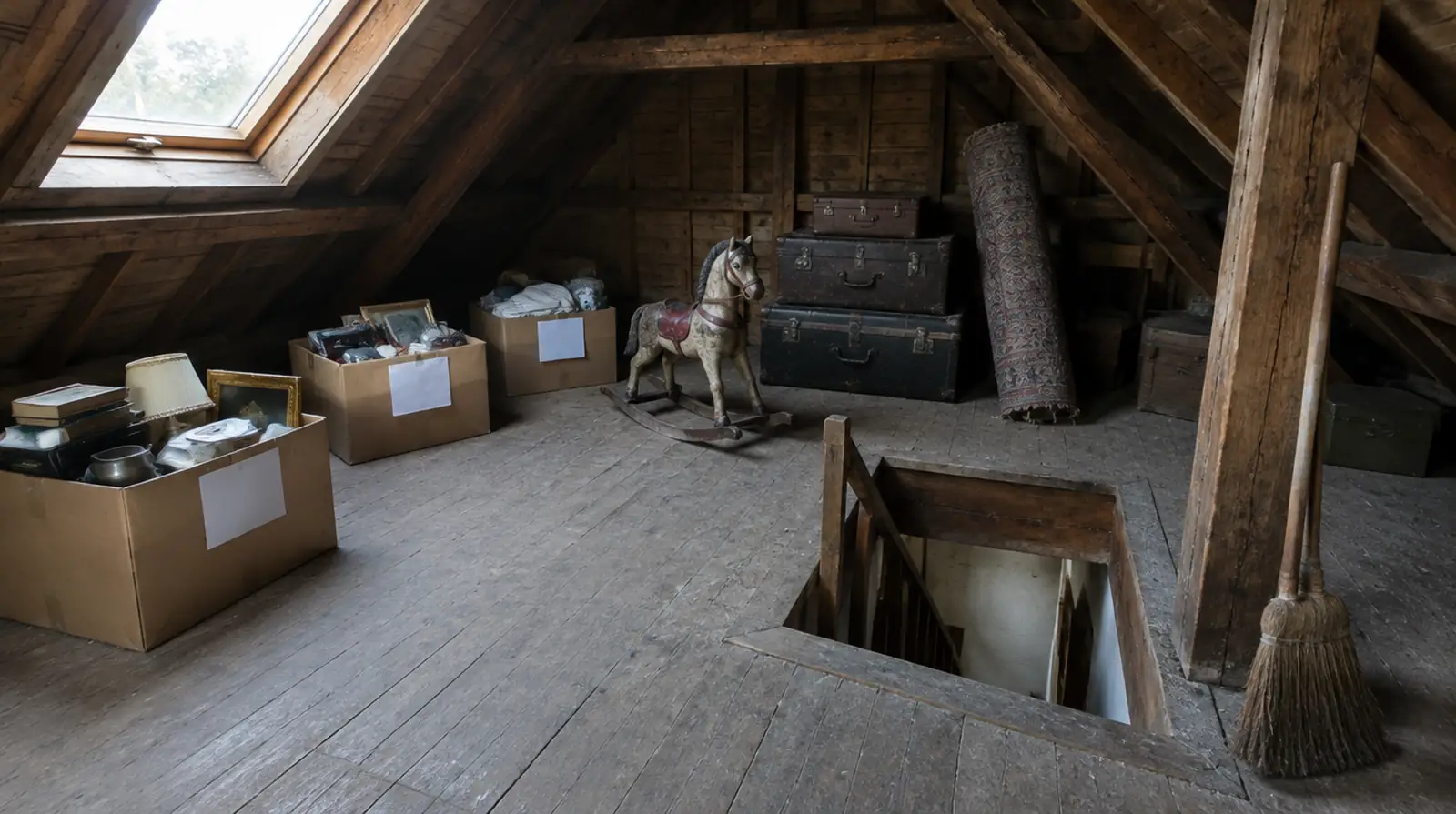 Een half leeggeruimde zolder onder houten balken, dozen in drie groepen, oude koffers en een hobbelpaard bij het trapgat
